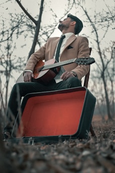 A man in a tan blazer and tie is sitting on a chair outdoors, holding a classical guitar. An open, empty guitar case lies on the ground nearby. The barren trees and dry leaves suggest a winter or autumn setting.
