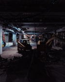 A dimly lit construction site with industrial machinery, including a skid-steer loader and an excavator, positioned among concrete columns and debris. Overhead lighting casts a cold, muted glow, creating long shadows across the cluttered ground.