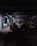 A dimly lit construction site with industrial machinery, including a skid-steer loader and an excavator, positioned among concrete columns and debris. Overhead lighting casts a cold, muted glow, creating long shadows across the cluttered ground.