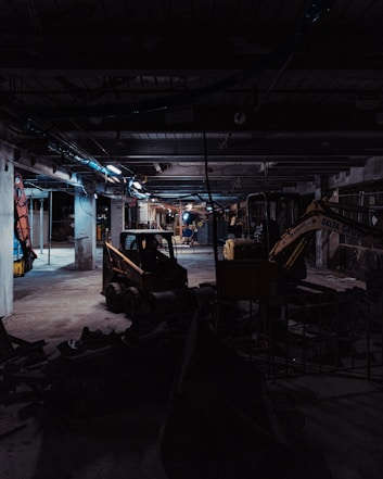 A dimly lit construction site with industrial machinery, including a skid-steer loader and an excavator, positioned among concrete columns and debris. Overhead lighting casts a cold, muted glow, creating long shadows across the cluttered ground.