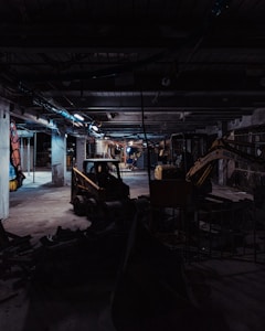 A dimly lit construction site with industrial machinery, including a skid-steer loader and an excavator, positioned among concrete columns and debris. Overhead lighting casts a cold, muted glow, creating long shadows across the cluttered ground.