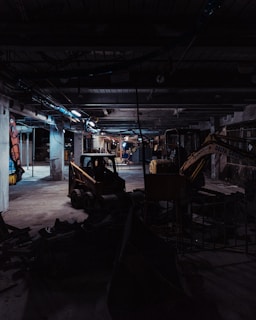 A dimly lit construction site with industrial machinery, including a skid-steer loader and an excavator, positioned among concrete columns and debris. Overhead lighting casts a cold, muted glow, creating long shadows across the cluttered ground.