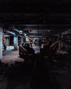 A dimly lit construction site with industrial machinery, including a skid-steer loader and an excavator, positioned among concrete columns and debris. Overhead lighting casts a cold, muted glow, creating long shadows across the cluttered ground.
