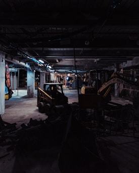 A dimly lit construction site with industrial machinery, including a skid-steer loader and an excavator, positioned among concrete columns and debris. Overhead lighting casts a cold, muted glow, creating long shadows across the cluttered ground.
