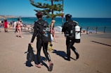 Two scuba divers in wetsuits are walking on a concrete pathway towards a sandy beach. They are carrying diving equipment, including tanks and fins. In the background, beachgoers can be seen either walking or lounging by the sea. A tree provides a bit of shade on the pathway, and the ocean is calm with a clear blue sky overhead.
