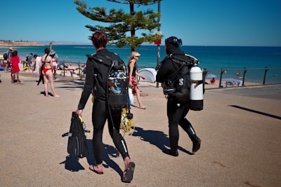 Two scuba divers in wetsuits are walking on a concrete pathway towards a sandy beach. They are carrying diving equipment, including tanks and fins. In the background, beachgoers can be seen either walking or lounging by the sea. A tree provides a bit of shade on the pathway, and the ocean is calm with a clear blue sky overhead.