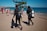 Two scuba divers in wetsuits are walking on a concrete pathway towards a sandy beach. They are carrying diving equipment, including tanks and fins. In the background, beachgoers can be seen either walking or lounging by the sea. A tree provides a bit of shade on the pathway, and the ocean is calm with a clear blue sky overhead.