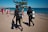 Two scuba divers in wetsuits are walking on a concrete pathway towards a sandy beach. They are carrying diving equipment, including tanks and fins. In the background, beachgoers can be seen either walking or lounging by the sea. A tree provides a bit of shade on the pathway, and the ocean is calm with a clear blue sky overhead.