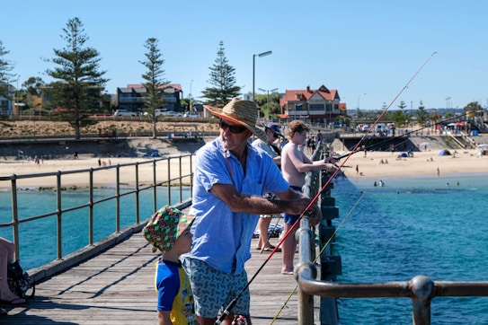 A man wearing a straw hat and sunglasses is fishing on a pier with a young child beside him. The weather is sunny, with a clear blue sky and several people in swimsuits on the pier and in the background on the shore. There are houses and trees lining the coast, and the water in the foreground is a vibrant blue-green.