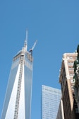 Skyscrapers dominate the scene, with a clear blue sky as the backdrop. The tallest building has cranes on top, indicating construction. Adjacent buildings feature modern glass facades and classic brick architecture.