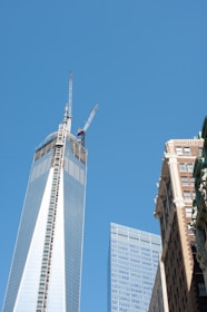 Skyscrapers dominate the scene, with a clear blue sky as the backdrop. The tallest building has cranes on top, indicating construction. Adjacent buildings feature modern glass facades and classic brick architecture.