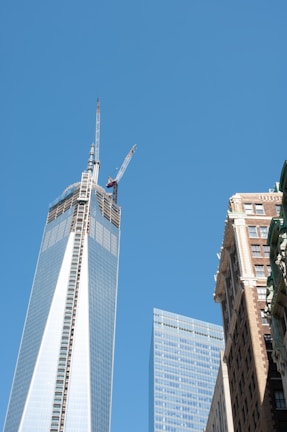 Skyscrapers dominate the scene, with a clear blue sky as the backdrop. The tallest building has cranes on top, indicating construction. Adjacent buildings feature modern glass facades and classic brick architecture.