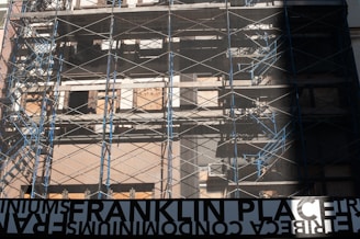 A partially constructed or renovated building surrounded by metal scaffolding. The structure has a raw, unfinished look with plywood covering some windows and metal beams crisscrossing the facade. Blue and silver tones dominate with the sunlight casting shadows. A large, reversed sign at the bottom reads 'FRANKLIN PLACE'.