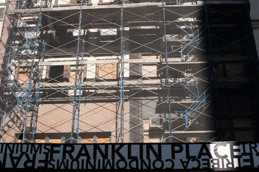 A partially constructed or renovated building surrounded by metal scaffolding. The structure has a raw, unfinished look with plywood covering some windows and metal beams crisscrossing the facade. Blue and silver tones dominate with the sunlight casting shadows. A large, reversed sign at the bottom reads 'FRANKLIN PLACE'.