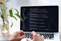 A person is typing on a laptop displaying a text editor with code. The scene is set on a light wooden table near a window, with a simple glass vase holding a few delicate flowers and green leaves.