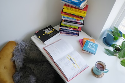 A well-lit study nook features a stack of books on a white desk. An open book lies in the center, surrounded by a smaller pile of colorful books and a mug filled with tea. A potted plant adds greenery, while a fluffy gray chair offers comfort.