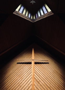 A wooden cross is mounted on a textured wooden wall beneath a sloping roof with an open skylight, which features stained glass panels of varying colors. Soft light filters through the window, illuminating the cross and creating a serene atmosphere.