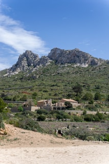 A rural landscape featuring a rocky mountain in the background, with clear blue skies above. In the foreground, there is a rustic farmhouse surrounded by lush greenery and terraced fields, giving a serene and peaceful countryside vibe.