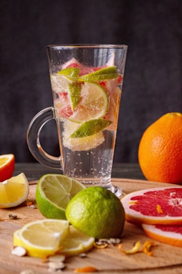 A tall glass filled with sparkling water and slices of various citrus fruits, including limes, grapefruits, and lemons, is placed on a wooden surface. Surrounding the glass are a variety of fresh citrus fruits, both whole and sliced. The backdrop is a dark, blurred surface, highlighting the colors of the fruits.