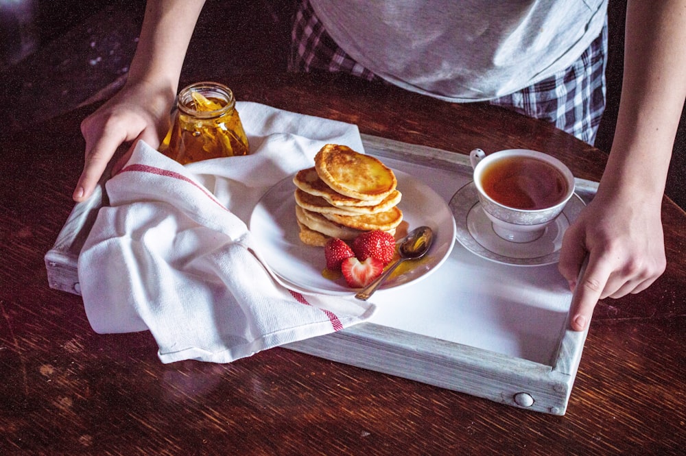 person holding white ceramic mug with coffee beside burger on white ceramic plate