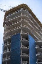 A high-rise building under construction with visible concrete floors and metal railings. The structure is partially covered with dark blue panels, and a construction crane is visible in the background.