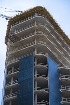 A high-rise building under construction with visible concrete floors and metal railings. The structure is partially covered with dark blue panels, and a construction crane is visible in the background.