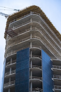 A high-rise building under construction with visible concrete floors and metal railings. The structure is partially covered with dark blue panels, and a construction crane is visible in the background.
