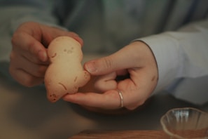 A pair of hands holds a small, cute cookie shaped like a character with simple facial features. The cookie appears to be freshly baked, with a light brown color. A wooden surface and a glass bowl are partially visible in the background.