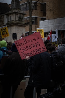 A protest scene with several people holding placards. The central sign reads 'Priti Patel is a serious annoyance.' Background signs include messages like 'Kill the Bill' and 'Black Lives Matter.' The setting appears to be a public area with an old stone monument, trees, and modern buildings in the background.