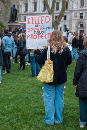A protest scene with people gathered on a grassy area, one person in the foreground is holding a sign that reads 'KILLED BY THE SYSTEM WE'RE TOLD TO PROTECT' written in red and black letters. The individual appears to have long hair and is wearing casual clothing, including a black top, blue jeans, and carrying a yellow tote bag. There are other protestors in the background, some holding signs as well, and a statue is visible in the park area.