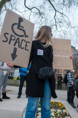 A woman holding two cardboard signs, one with a wheelchair symbol and the word 'SPACE' and the other with the phrase 'NO MEANS NO.' She stands in an outdoor setting on a overcast day, surrounded by a few people, some of whom are wearing masks. In the background are trees without leaves and some buildings.
