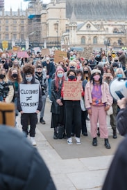 A large crowd of people, mostly wearing masks, are gathered together holding various protest signs. They are standing in front of an older building with Gothic architecture. The scene suggests a protest or rally. The signs include messages about consent and safety.