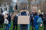 A large group of people gathered outdoors for a protest. Many individuals hold signs displaying messages related to rights and opposition to government policies. Participants are largely dressed in casual clothing, with some wearing backpacks. The setting appears to be an urban area, with historic buildings visible in the background and a statue amongst the crowd.