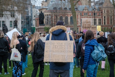 A large group of people gathered outdoors for a protest. Many individuals hold signs displaying messages related to rights and opposition to government policies. Participants are largely dressed in casual clothing, with some wearing backpacks. The setting appears to be an urban area, with historic buildings visible in the background and a statue amongst the crowd.