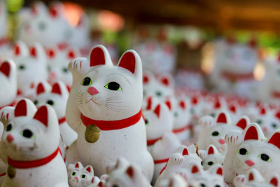 A collection of ceramic cat figurines featuring white bodies, red ears, and collars. The cats have one paw raised as if waving and are adorned with green eyes and small golden bells hanging from their necks.