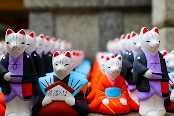 A collection of colorful painted fox figurines dressed in traditional Japanese clothing. They have different colored kimonos, predominantly with black, red, blue, and purple colors. Each figurine has a distinctive expression, some holding objects such as a folded paper or book with Japanese characters.