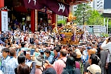 A large crowd gathers around a decorated portable shrine being carried by several people, surrounded by many onlookers. The scene appears festive, with people taking photos and wearing traditional clothing. The event is taking place outdoors near a temple, with greenery and city buildings visible in the background.