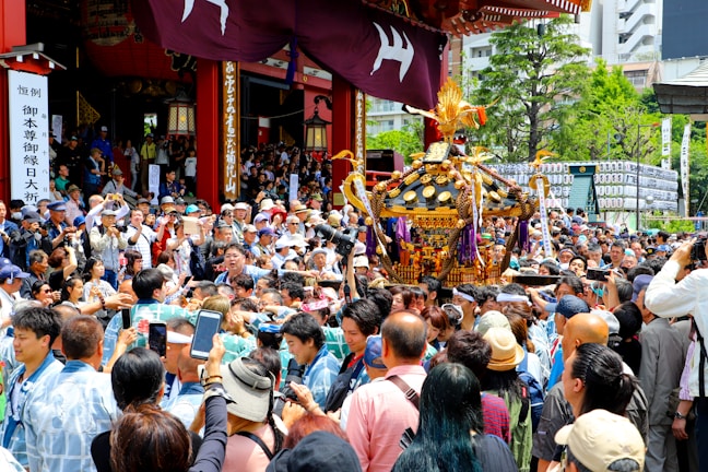 A large crowd gathers around a decorated portable shrine being carried by several people, surrounded by many onlookers. The scene appears festive, with people taking photos and wearing traditional clothing. The event is taking place outdoors near a temple, with greenery and city buildings visible in the background.