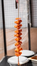 A skewer holds several pieces of marinated paneer interspersed with slices of onions and tomatoes. The ingredients are coated in a rich, spicy orange marinade. The skewer is held vertically, and there are blurred kitchen containers in the background.