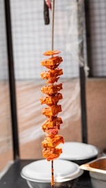 A skewer holds several pieces of marinated paneer interspersed with slices of onions and tomatoes. The ingredients are coated in a rich, spicy orange marinade. The skewer is held vertically, and there are blurred kitchen containers in the background.