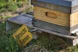 A wooden beehive rests on a wooden platform surrounded by grass. A yellow caution sign with black text warns of bees. The scene is set outdoors, likely in a rural or natural area.