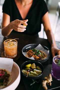 A person is sitting at a table with various dishes and drinks. There is a taco with shredded meat and fresh herbs, a bowl of lime wedges, and two colorful cocktails, one orange and one purple. The person is holding a slice of lime, about to squeeze it. The atmosphere seems casual and relaxed.