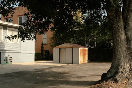 A small, rusted metal shed is situated on a concrete surface near a large tree and adjacent to a residential building with siding and windows. The area is shaded by tree branches, creating a quiet and somewhat neglected atmosphere.