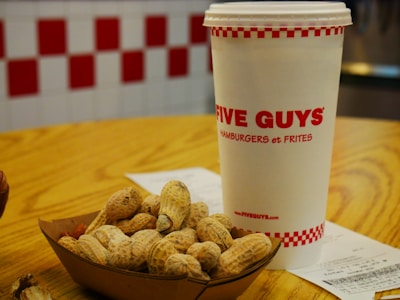 A takeaway cup with the logo 'Five Guys', placed on a wooden table next to a cardboard tray filled with in-shell peanuts. A receipt is partially visible on the table. The background features a wall with a red and white checkered pattern.