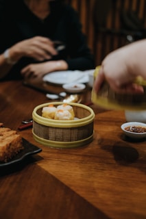 A traditionally styled bamboo steamer basket contains two pieces of dim sum placed on a wooden table. Nearby, a hand reaches for the basket, and another person is holding chopsticks. Plates and a small bowl of sauce are on the table, creating a setting typical of an Asian dining experience.