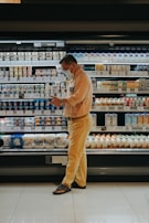 A person wearing a mask stands in front of a grocery store's refrigerated section, seemingly focused on their phone. The shelves are stocked with various dairy products and beverages, each labeled with price tags. The person is dressed in a beige shirt and yellow pants, wearing sandals. The lighting is warm, highlighting the products.