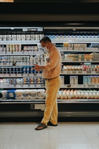 A person wearing a mask stands in front of a grocery store's refrigerated section, seemingly focused on their phone. The shelves are stocked with various dairy products and beverages, each labeled with price tags. The person is dressed in a beige shirt and yellow pants, wearing sandals. The lighting is warm, highlighting the products.