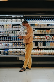 A person wearing a mask stands in front of a grocery store's refrigerated section, seemingly focused on their phone. The shelves are stocked with various dairy products and beverages, each labeled with price tags. The person is dressed in a beige shirt and yellow pants, wearing sandals. The lighting is warm, highlighting the products.