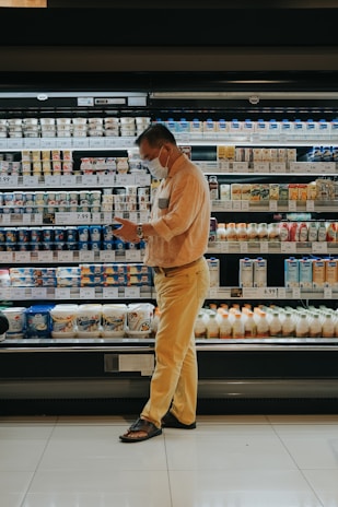 A person wearing a mask stands in front of a grocery store's refrigerated section, seemingly focused on their phone. The shelves are stocked with various dairy products and beverages, each labeled with price tags. The person is dressed in a beige shirt and yellow pants, wearing sandals. The lighting is warm, highlighting the products.