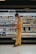 A person wearing a mask stands in front of a grocery store's refrigerated section, seemingly focused on their phone. The shelves are stocked with various dairy products and beverages, each labeled with price tags. The person is dressed in a beige shirt and yellow pants, wearing sandals. The lighting is warm, highlighting the products.